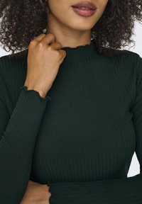 Ciemnozielona, prążkowana bluzka z długim rękawem o wysokim dekolcie oraz falbaniastych brzegach mankietów. Gładka faktura i dopasowany krój.