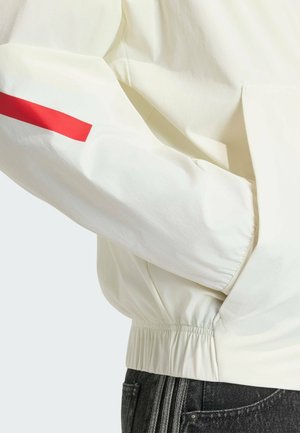 Manica di giacca bianca con una striscia rossa sopra il polsino elastico, mano nella tasca laterale, abbinata a jeans di denim scuro.