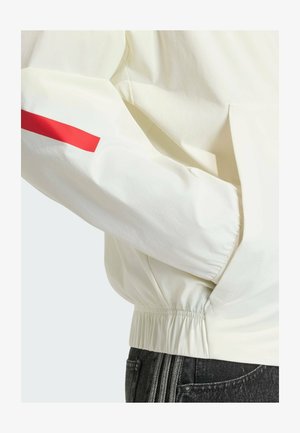 Manica di giacca bianca con una striscia rossa sopra il polsino elastico, mano nella tasca laterale, abbinata a jeans di denim scuro.