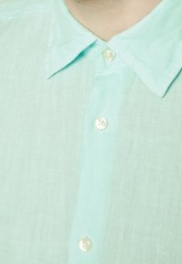 Jasno miętowy lniany ś-shirt z zaostrzonym kołnierzykiem, z białymi plastikowymi guzikami i teksturowaną powierzchnią materiału.