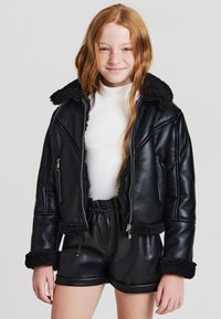 Giacca di pelle nera con colletto e polsini in shearling sintetico, chiusura con zip, design corto. Indossata sopra un dolcevita bianco e pantaloni corti neri.