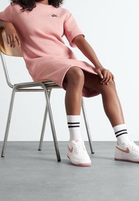 Růžová mikinová šaty Nike, střední délky. Bílé tenisky s růžovými akcenty, spárované s bílými ponožkami s černými pruhy. Jednoduchý design.