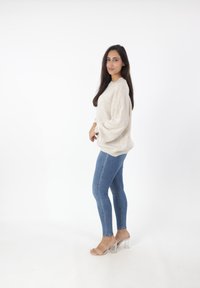 Hellbeige übergroßer Pullover mit weiten Ärmeln, kombiniert mit blauen Skinny Jeans und klaren Sandalen mit Absatz, vor einem schlichten weißen Hintergrund.