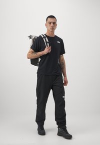 Camiseta preta e calças com o logo da The North Face, mochila cinza e preta, sapatos robustos, em pé contra um fundo branco liso.
