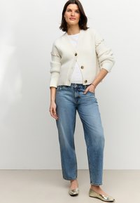 Krém színű kötött kardigán bordázott mintával és három gombbal, világoskék egyenes szárú farmerrel és fémes lapos cipővel párosítva.