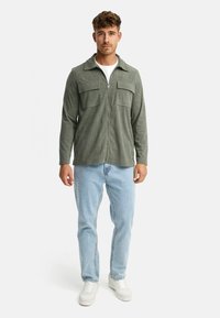 Mand iført olivengrønne lynlås corduroy skjorte over hvid t-shirt, lyseblå jeans og hvide sneakers, stående mod en ensfarvet baggrund.