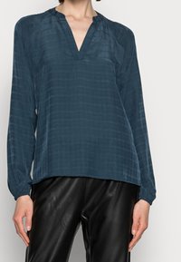 Blusa lunga a maniche con scollo a V di un verde scuro, in tessuto testurizzato, con maniche leggermente arricciate, abbinata a pantaloni di pelle nera.