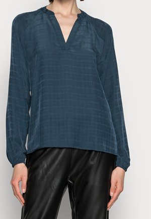 Blusa lunga a maniche con scollo a V di un verde scuro, in tessuto testurizzato, con maniche leggermente arricciate, abbinata a pantaloni di pelle nera.