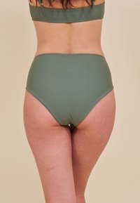 Kobieta widziana z tyłu, ubrana w oliwkowe, wysokie majtki bikini oraz pasujący top bikini na jednolitym beżowym tle.