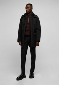 Lange zwarte parka met een rits en knoopsluiting, gedragen over een donkerrode sweater en zwarte broek, gecombineerd met zwarte chunky schoenen.