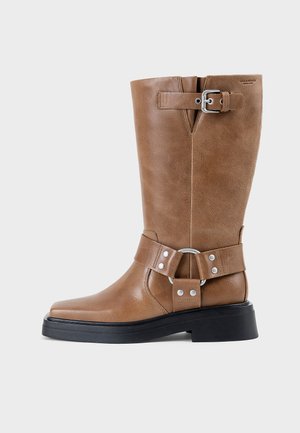 Bota de cuero marrón hasta la mitad de la pantorrilla con suela negra, hebilla plateada en la parte superior y remaches decorativos plateados alrededor de la correa del tobillo.