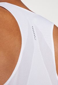 Hvit atletisk tank topp med mesh tekstur, scoop rygg og et reflekterende logo. Teksturert linjer går diagonal over stoffet.