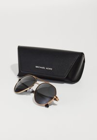 Óculos de sol Michael Kors com uma armação de metal dourado, lentes escuras e uma caixa de couro preto com fecho de pressão e logótipo em relevo.
