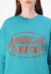 Teal sweatshirt med en cirkulär logotyp som har texten "AMERICAN VINTAGE" i rosaguld metallictryck. Mjuk, texturerad tyg med rundad hals.
