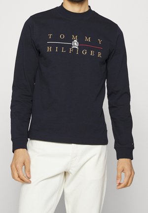 Hombre vestido con una camisa de manga larga negra con el texto y emblema dorados "TOMMY HILFIGER", combinada con pantalones blancos.