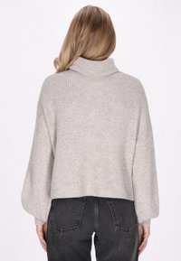 Hellgrauer Strickpullover mit hohem Kragen, geripptem Muster und überschnittenen Schultern, kombiniert mit dunklen, hochtaillierten Jeans.