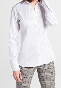 Chemise blanche à manches longues avec col pointu, tissu en coton lisse et boutons en perle. Portée avec un pantalon à motifs à carreaux.