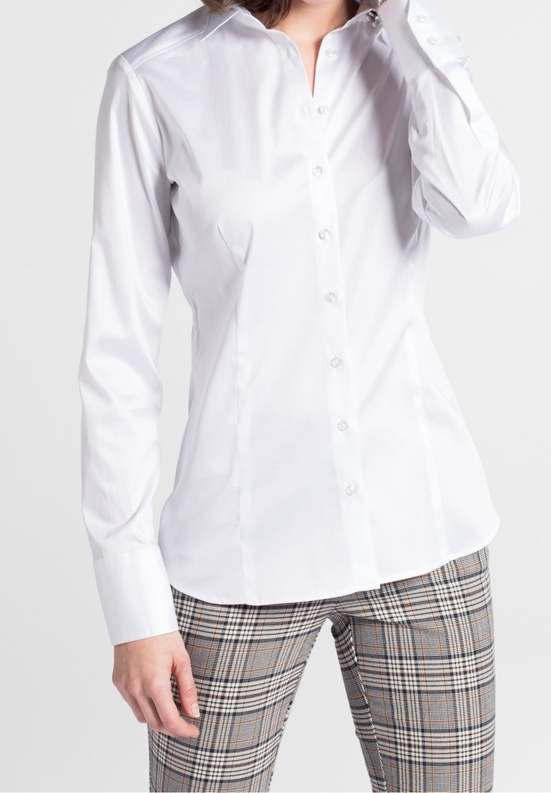 Chemise blanche à manches longues avec col pointu, tissu en coton lisse et boutons en perle. Portée avec un pantalon à motifs à carreaux.