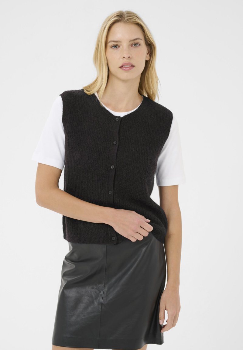 Gilet nero lavorato a maglia con scollatura rotonda e bottoni davanti, indossato sopra una camicia bianca a maniche corte. Combinato con una gonna in pelle nera.