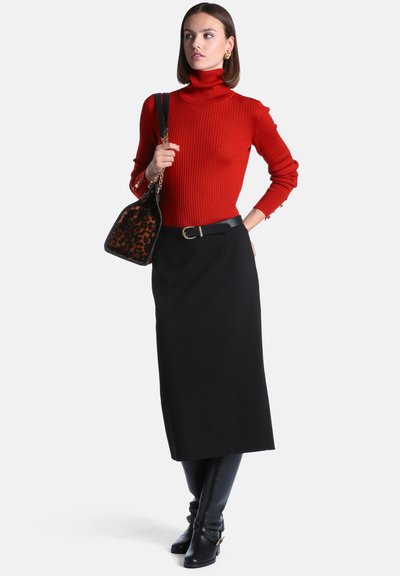 Femme portant un pull à col roulé côtelé rouge, une jupe midi noire, une ceinture noire, des bottes noires montantes aux genoux, et un sac à main imprimé léopard sur l'épaule, regardant sur le côté.
