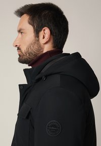 Czarna kurtka z kapturem z zamkiem, z teksturowanym bordowym kołnierzem i okrągłą naszywką marki na lewym rękawie. Prosty design, praktyczna.