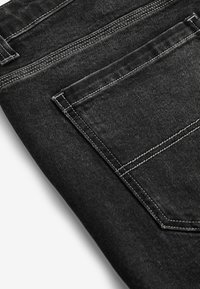 Zwart denim jeans met een opvallende textuur, voorzien van witte stiksels en een achterzak. De stof vertoont een lichte ribbelstructuur.