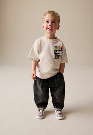 Beżowy bawełniany t-shirt z logo Marvel i grafiką postaci, zestawiony z ciemnymi, oversize'owymi spodniami jeansowymi oraz szarymi trampkami z dekoracyjnymi akcentami.