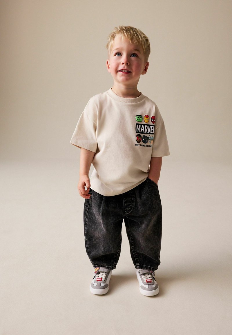 Bež bombažna majica z logotipom Marvel in grafikami likov, kombinirana s temnimi prevelikimi denim hlačami in sivimi supergami z okrasnimi poudarki.