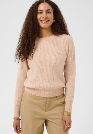 Femme aux longs cheveux bouclés portant un pull côtelé rose pâle et un pantalon beige, souriante devant un fond uni.