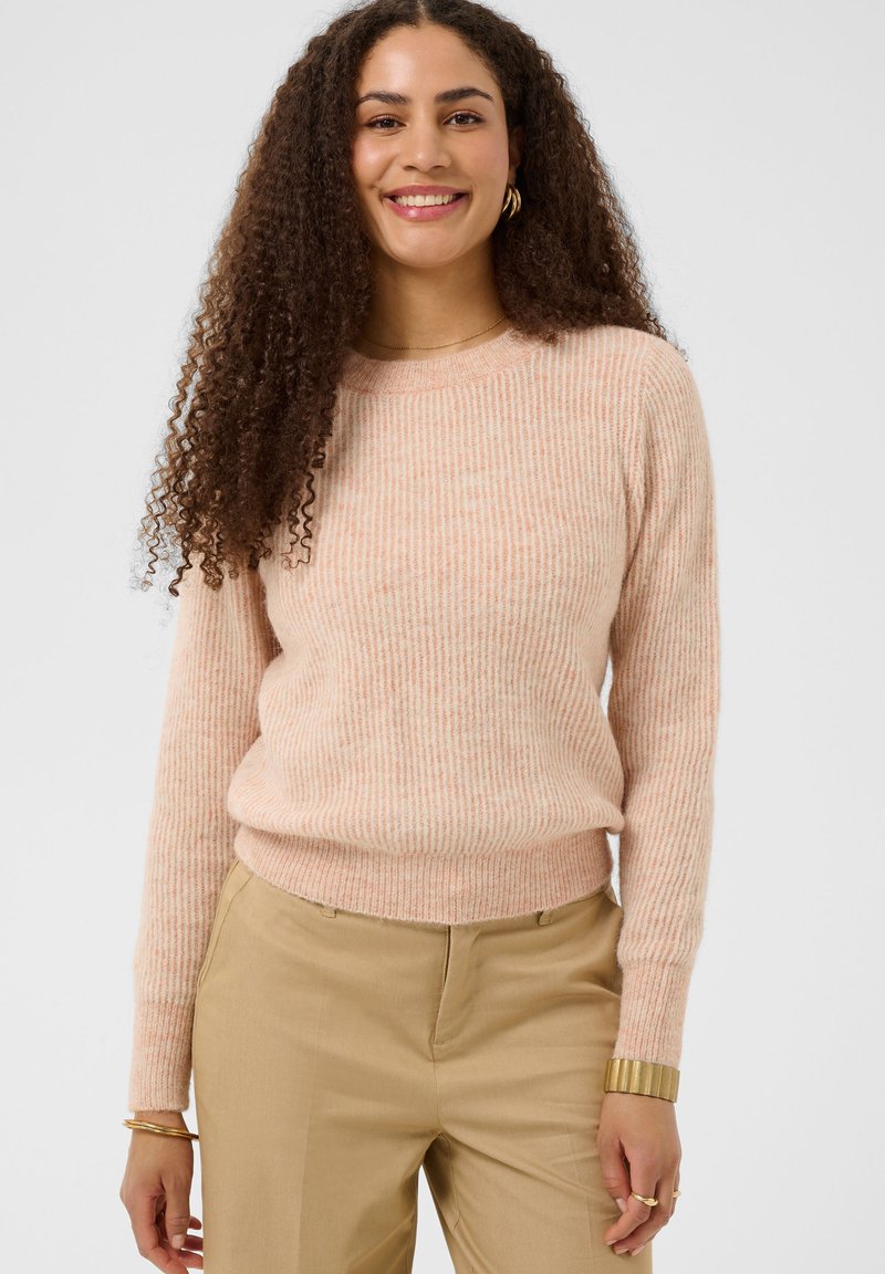 Femme aux longs cheveux bouclés portant un pull côtelé rose pâle et un pantalon beige, souriante devant un fond uni.