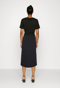 Donna con i capelli raccolti in un chignon, che indossa una maglietta nera a maniche corte, una gonna midi blu navy e sandali con tacco a blocco beige, girata di spalle su sfondo bianco.