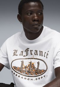 Hvid bomulds t-shirt med et grafisk tryk med brune og orange elementer, der viser tekst og et abstrakt ildagtigt design.