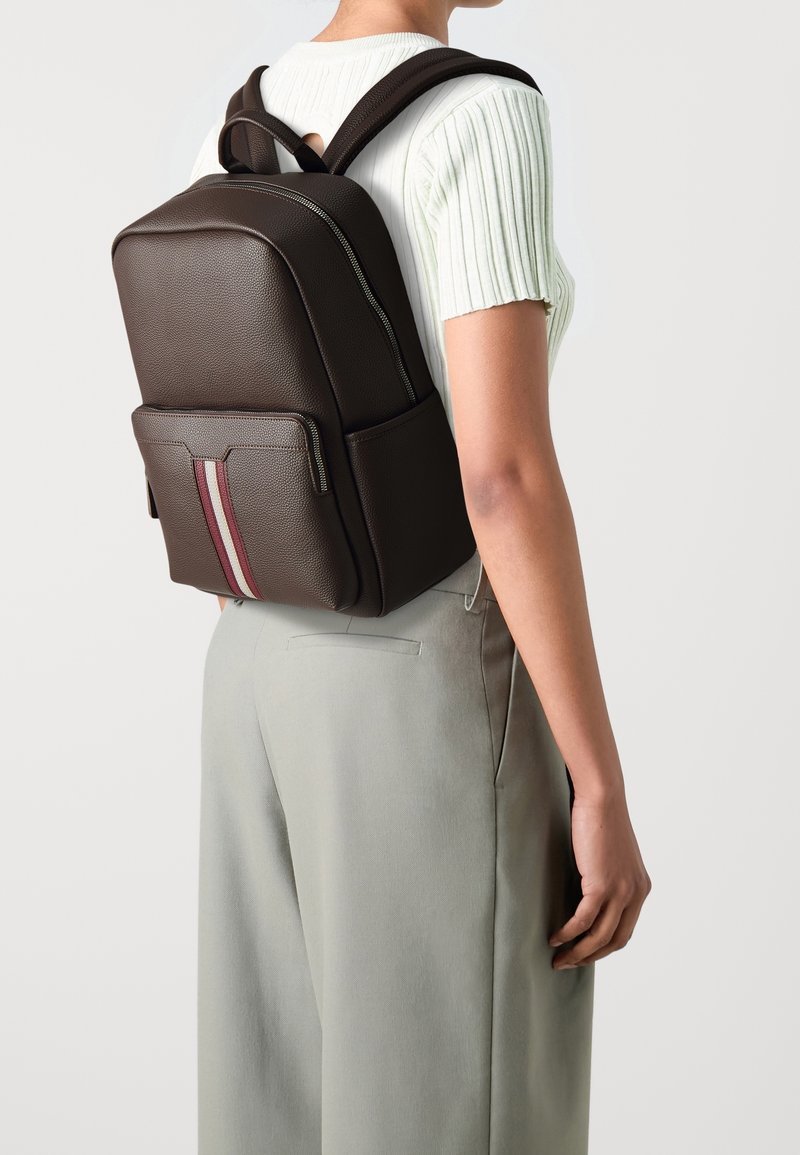 Sac à dos en cuir marron avec une texture lisse, rayures verticales rouges et blanches, deux poches latérales et bretelles rembourrées.