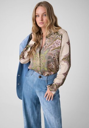 Jonge vrouw met lang blond haar, draagt een blouse met lange mouwen en patroon en lichtblauwe high-waisted broek, houdt een blauw jasje over haar schouder.