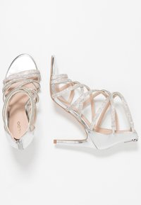 Silberne High-Heelsandalen mit überkreuzten Riemen, verziert mit Strasssteinen, in metallischer Optik und mit einem spitzen Zehenbereich.