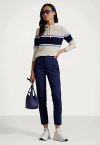 Mulher a usar óculos de sol azuis, uma sweatshirt listrada em branco e azul-marinho, calças azul-marinho, ténis brancos, a segurar uma mala azul-marinho, a posar contra um fundo simples.