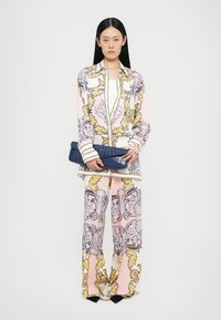 Zijden outfit met een roze blouse met decoratieve gele en grijze patronen, gecombineerd met bijpassende wijde broek. Een blauwe clutch wordt in de hand gehouden.