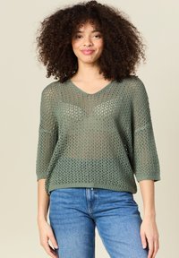 Grüner gestrickter Pullover mit V-Ausschnitt, 3/4-Ärmeln und offenem Webmuster, kombiniert mit blauen Jeans. Strukturiertes und leichtes Material.