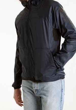 Homme portant une veste noire zippée avec poches et un jean bleu clair, debout avec une main dans une poche sur un fond uni.