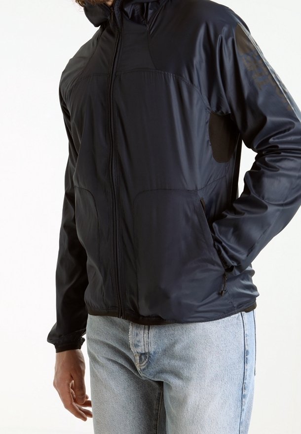 Homme portant une veste noire zippée avec poches et un jean bleu clair, debout avec une main dans une poche sur un fond uni.