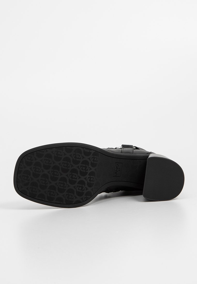 Zapato negro de tacón con una parte superior de cuero suave, acento de hebilla y una suela de goma texturizada con un patrón circular.