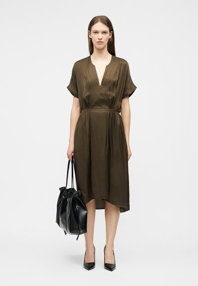 Femme portant une robe midi marron à manches courtes avec un décolleté profond en V, tenant un grand sac à main en cuir noir, et chaussée de talons pointus noirs.