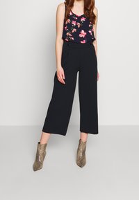 Pantalon large noir à jambes larges avec deux boutons sur le côté, associé à un débardeur bleu marine orné de motifs floraux roses. Bottines chevilles à imprimé serpent texturé.