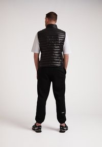 Gilet nero in stile piumino con sezioni a righe orizzontali, indossato sopra una maglietta bianca. Abbinato a pantaloni jogger neri e scarpe da ginnastica, visto da dietro.