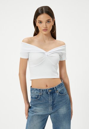 Witte geribbelde crop top met een off-shoulder ontwerp en knoopdetail aan de voorkant, gecombineerd met hoge blauwe spijkerbroeken.