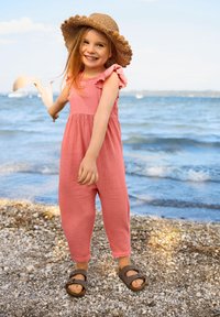 Rosa Baumwollspielanzug mit Rü schenärmeln, lockerer Passform und elastischen Knöcheln. Ein gewebter Strohhut und braune Sandalen runden das Outfit ab.