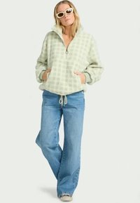 Grön rutig fleecepullover med halv dragkedja och framfickor, ihop med ljusblå utsvängda jeans och grå sneakers.