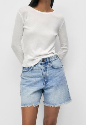 Vrouw met een witte top met lange mouwen en hoge taille lichtblauwe denim shorts met gerafelde zomen tegen een witte achtergrond.