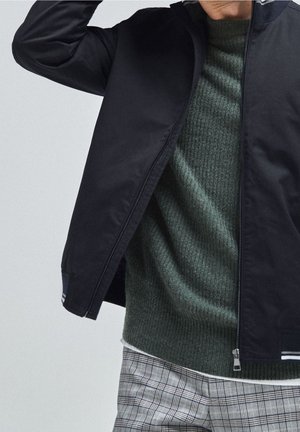 Personne portant une veste bleu foncé à fermeture éclair sur un pull vert en maille et un pantalon gris à carreaux, le bras levé près de la tête.