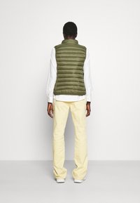 Grön pufferväst med hög krage, lätt vadderad textur, bärs över en vit långärmad skjorta, matchad med gula byxor och vita skor.